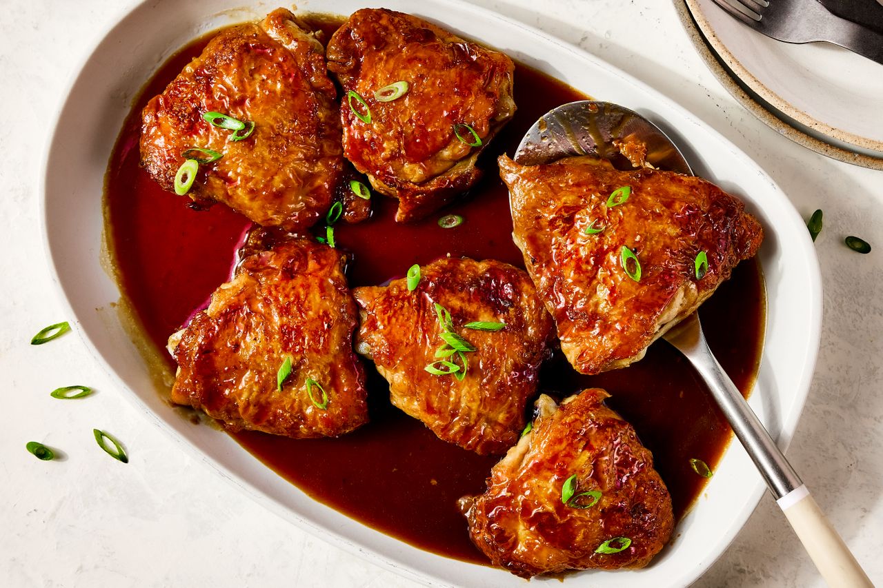 Mahogany chicken thighs in a sauce garnished with thinly sliced scallions in a baking dish with serving utensils nearby