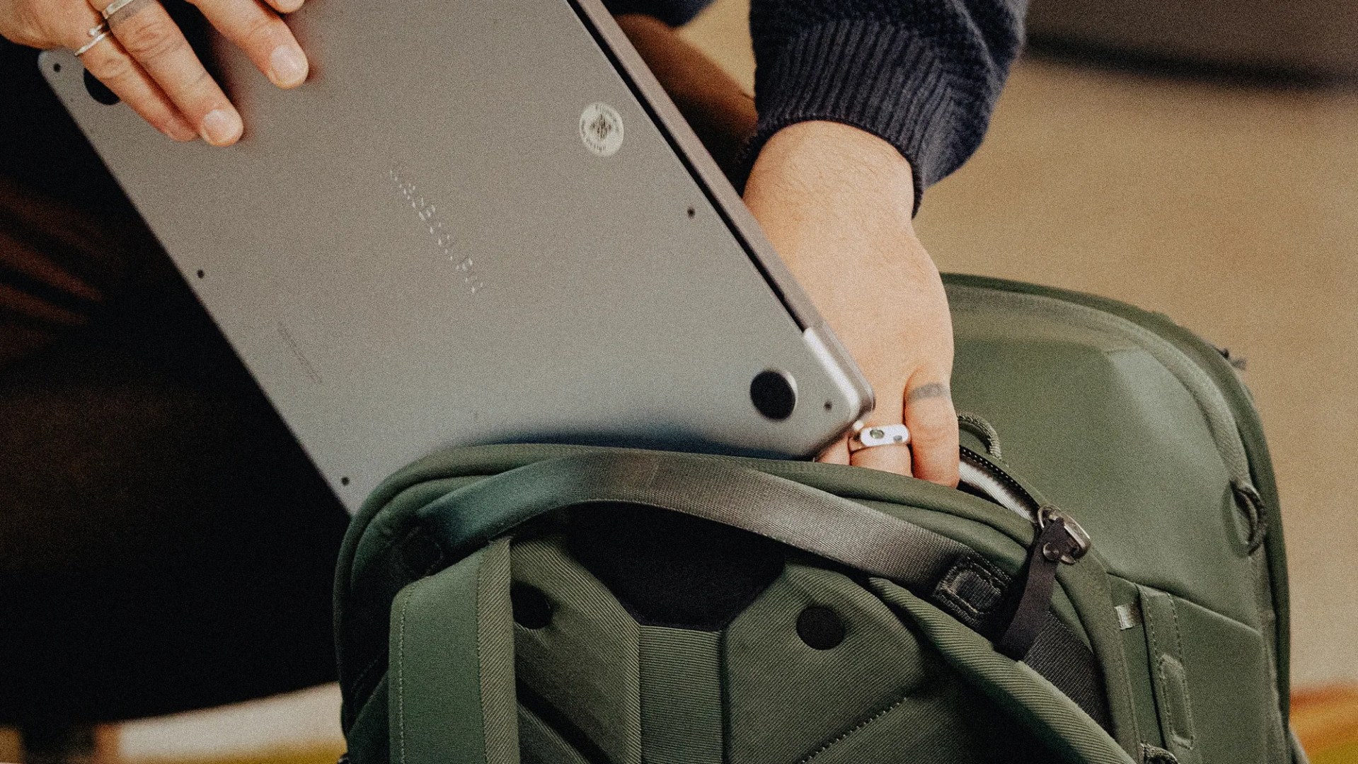 Person placing a silver laptop into an open dark green backpack with padded compartments.