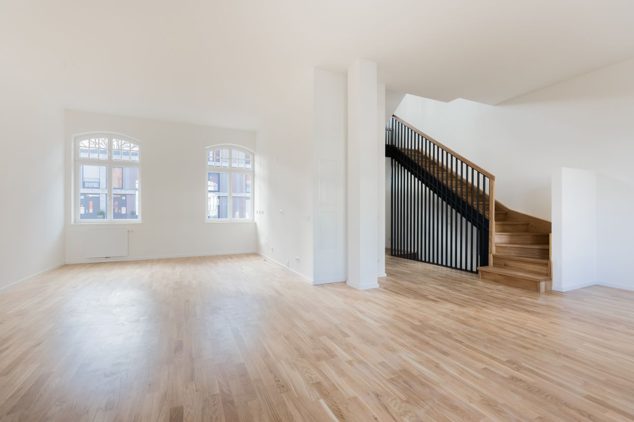 Large empty interior space with wooden flooring large windows and a staircase leading upward on the right side