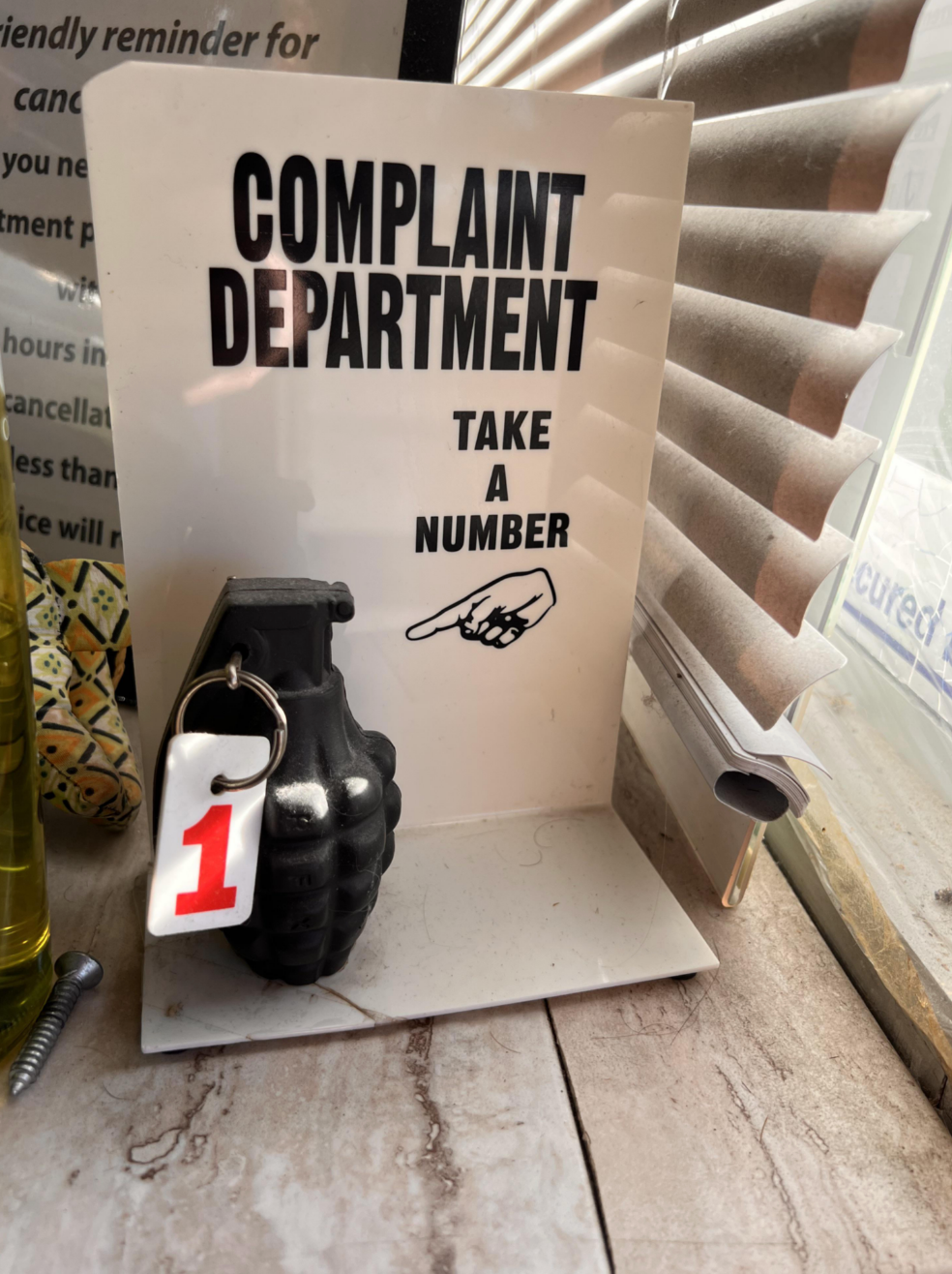 A humorous desk sign labeled "COMPLAINT DEPARTMENT" with a grenade and a "Take a Number" tag attached to a pin