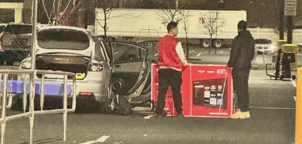 Two individuals stand by a car in a parking lot at night, unloading a large flat-screen TV box. A hotel building is visible in the background