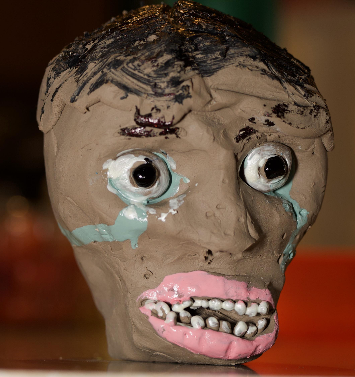 Clay sculpture of a human head with exaggerated features. The eyes are wide, and the mouth reveals uneven teeth