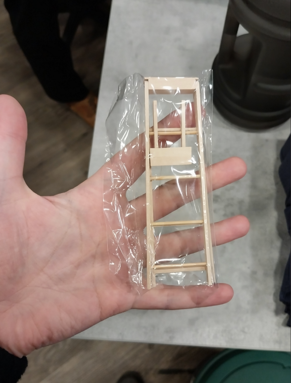 Hand holding a small wooden ladder encased in plastic wrap