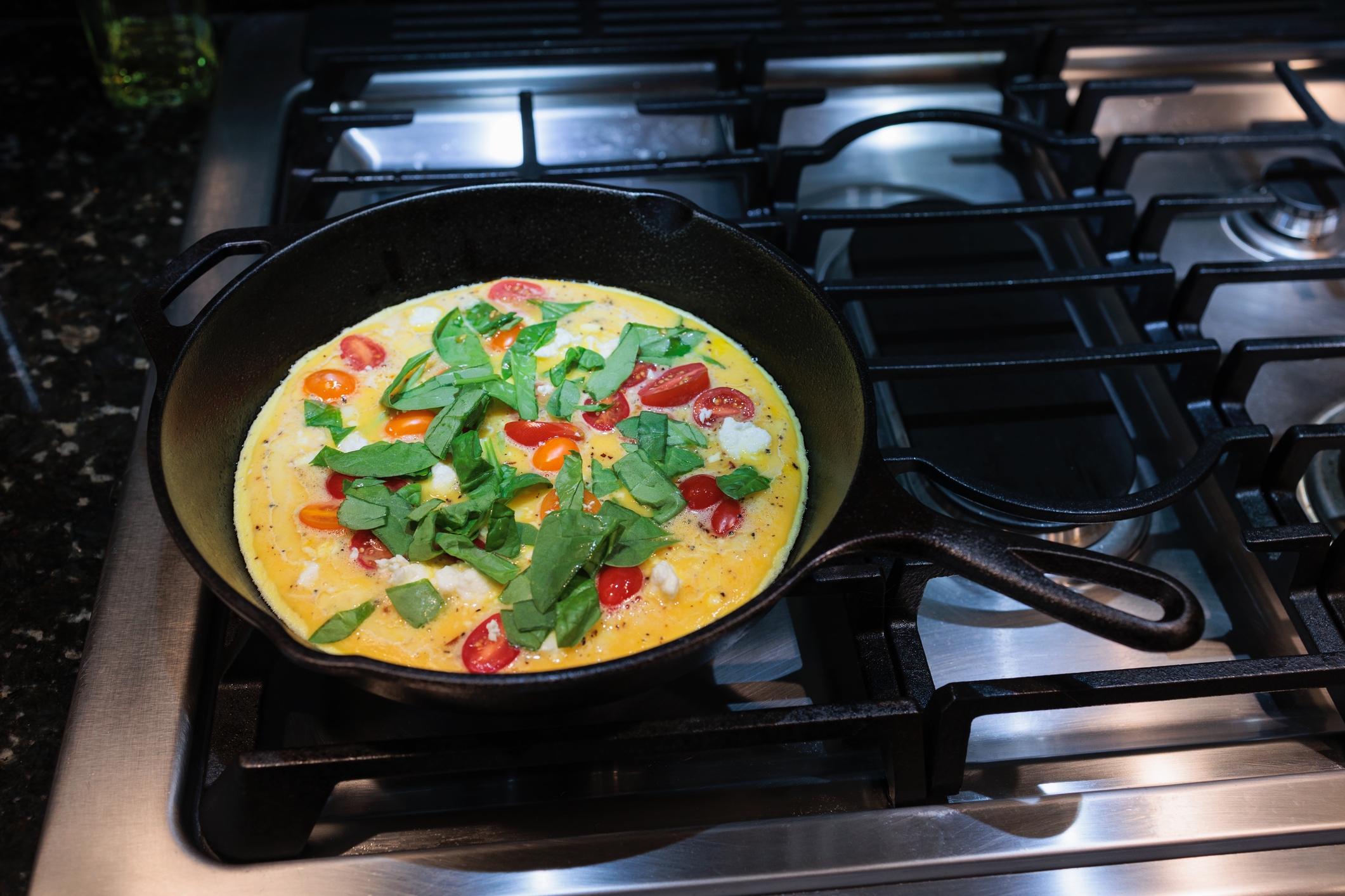 Omelet in a skillet on a stove, topped with fresh spinach and cherry tomatoes