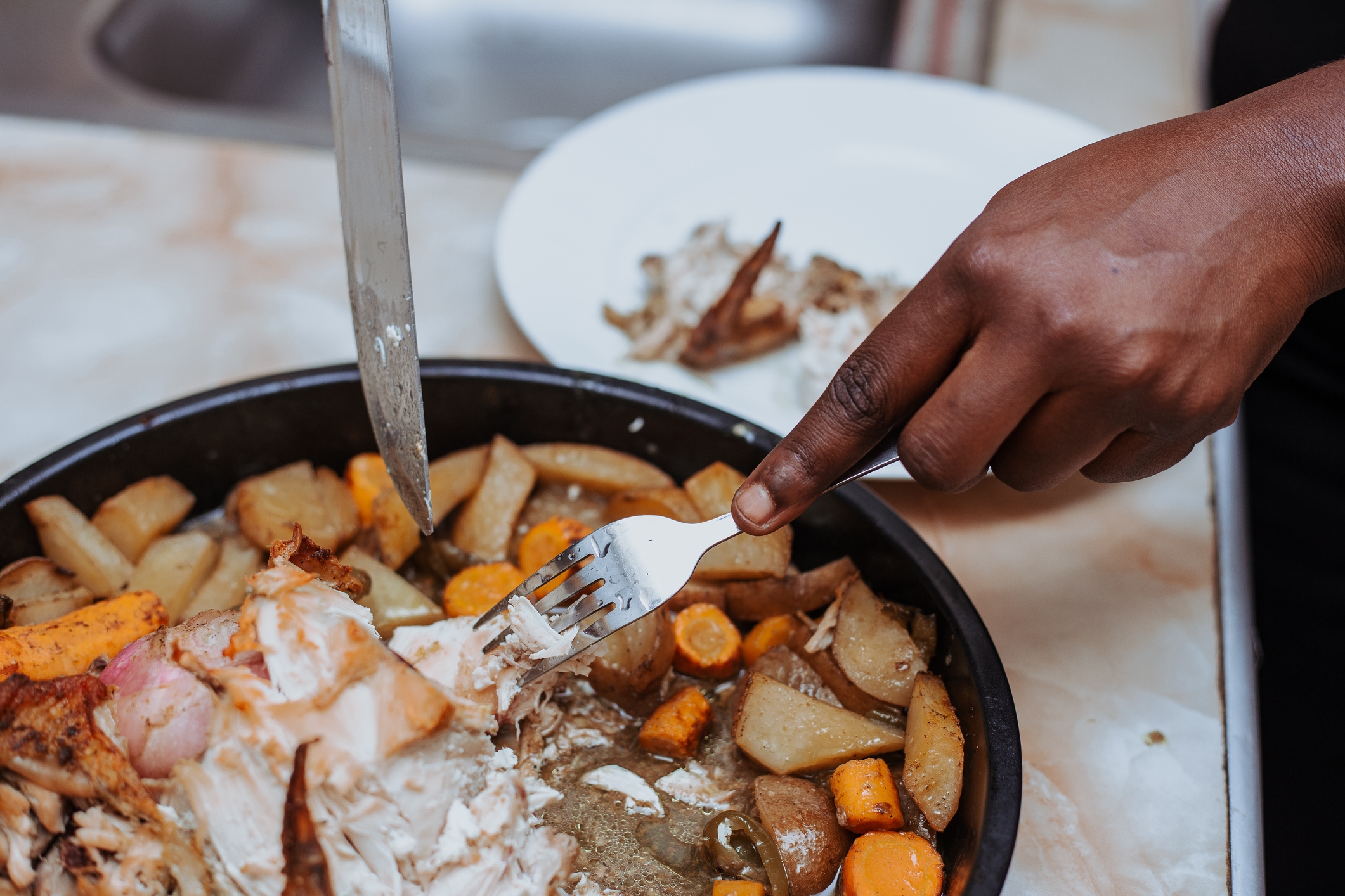 Person serving cooked chicken and vegetables from a pan onto a plate using a fork and knife