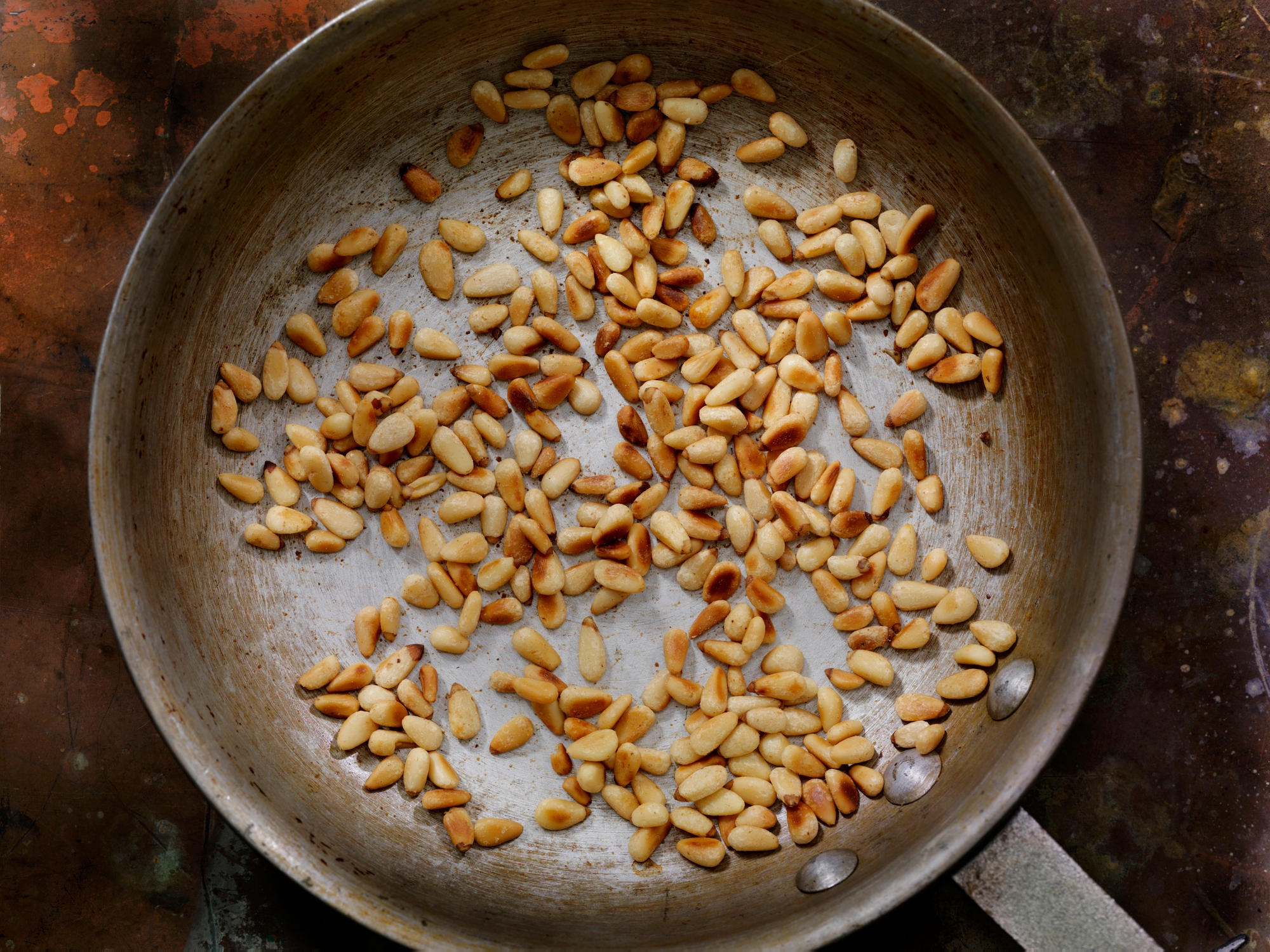 Toasted pine nuts scattered in a frying pan, some slightly browned