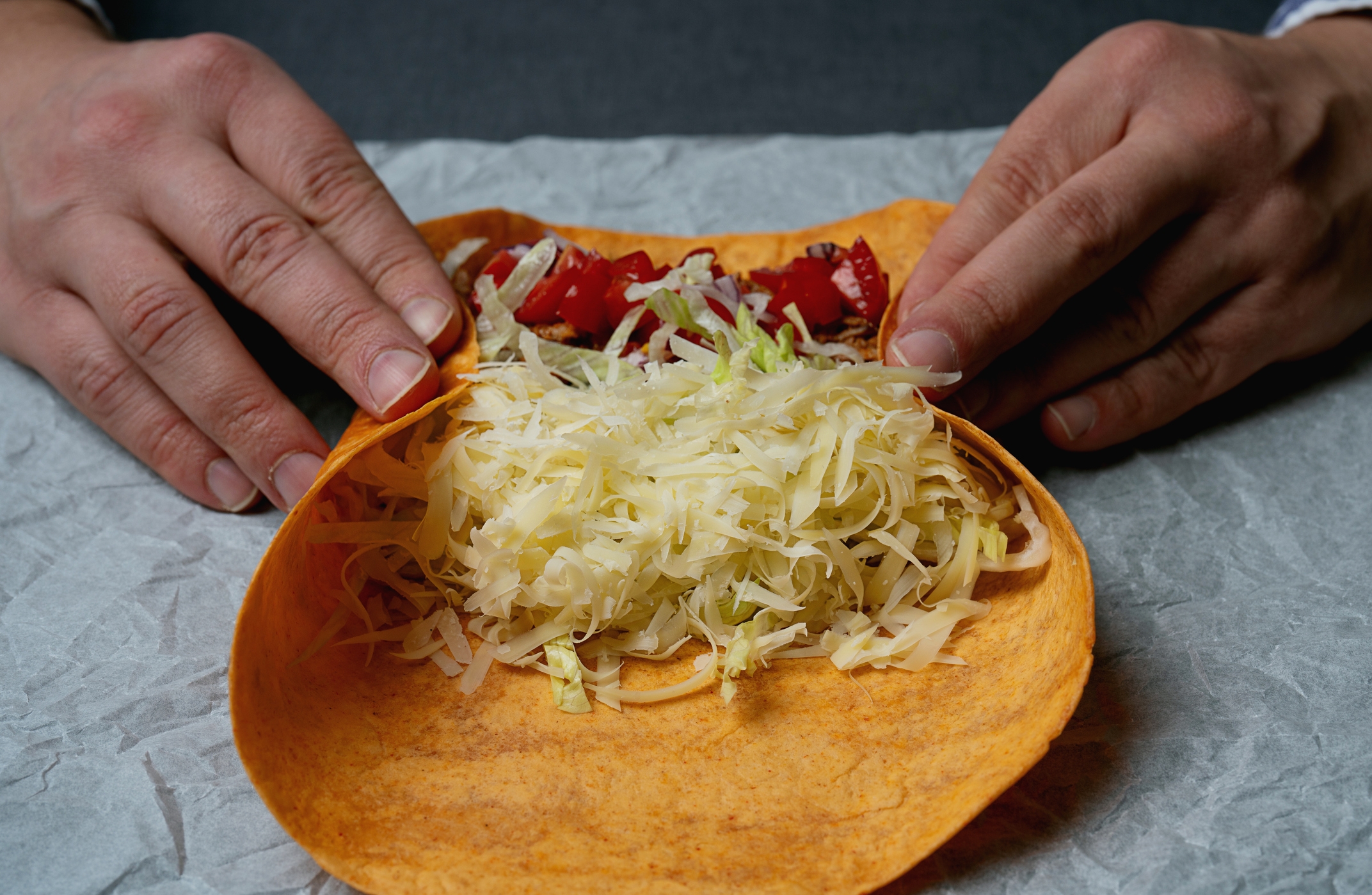 Person preparing to fold a tortilla filled with shredded cheese, lettuce, and chopped tomatoes