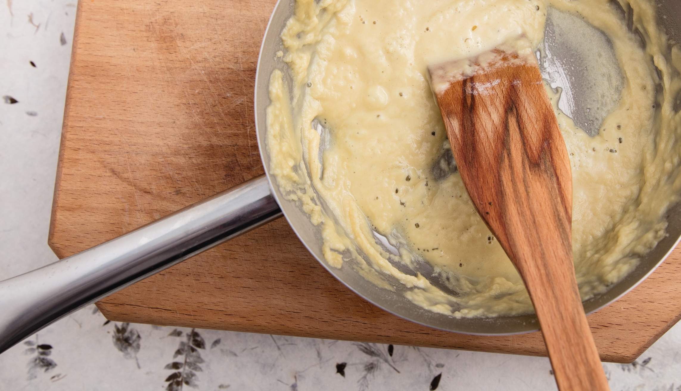 Pan with creamy sauce being stirred by a wooden spatula, resting on a wooden cutting board
