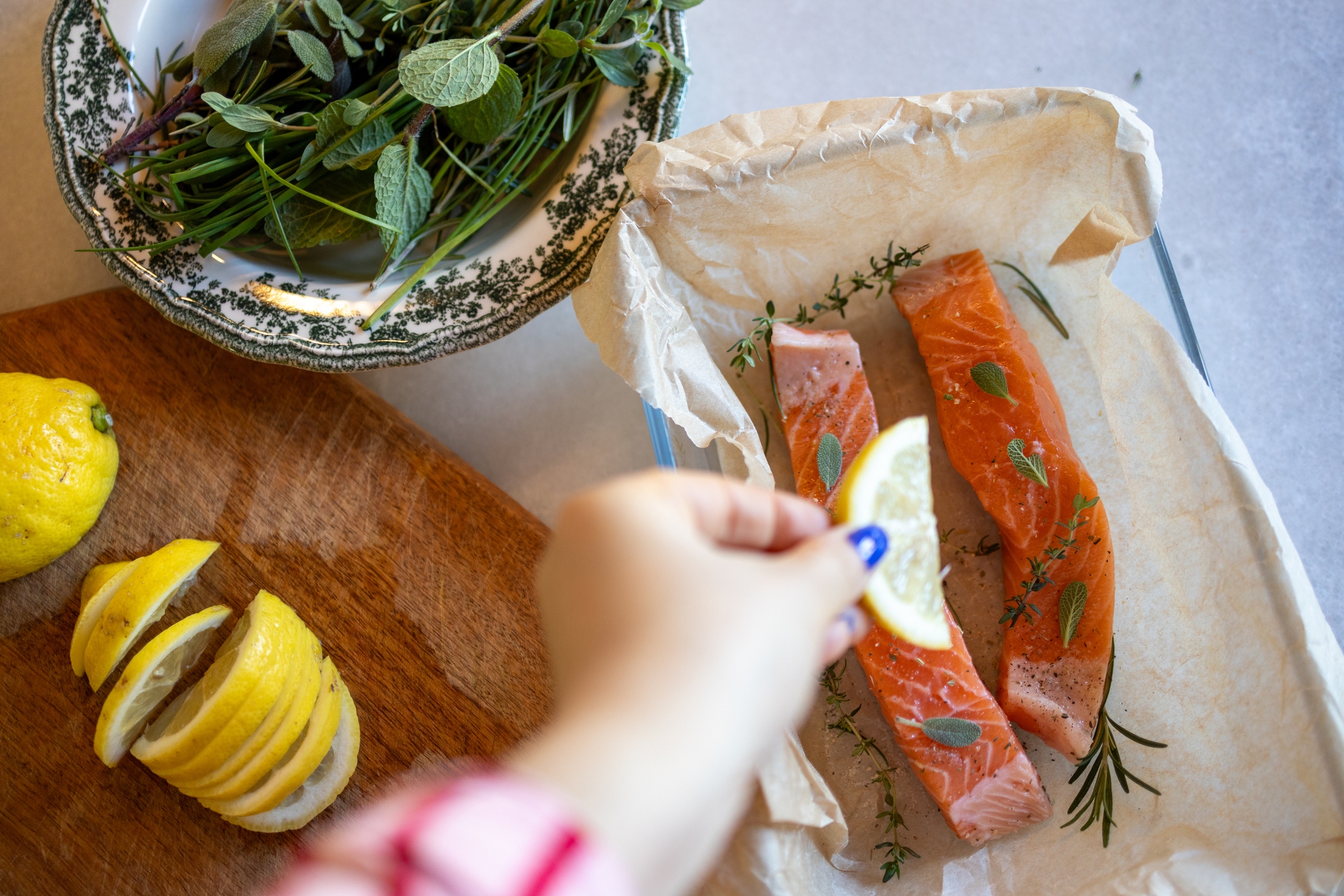Hand placing a lemon slice on uncooked salmon fillets on parchment, surrounded by fresh herbs and sliced lemons