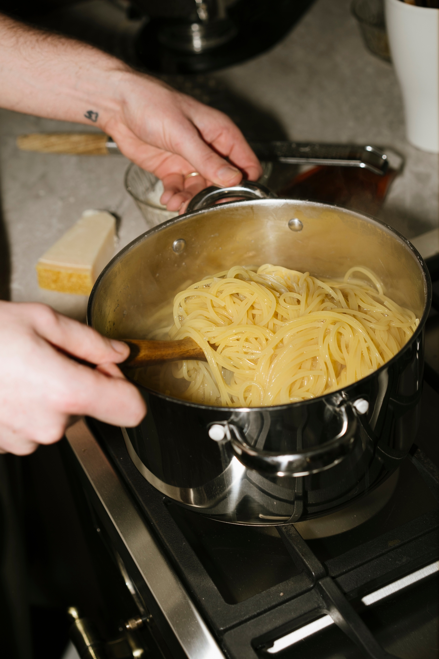 Person stirring spaghetti in a pot on a stove, with cheese and sauce ingredients nearby