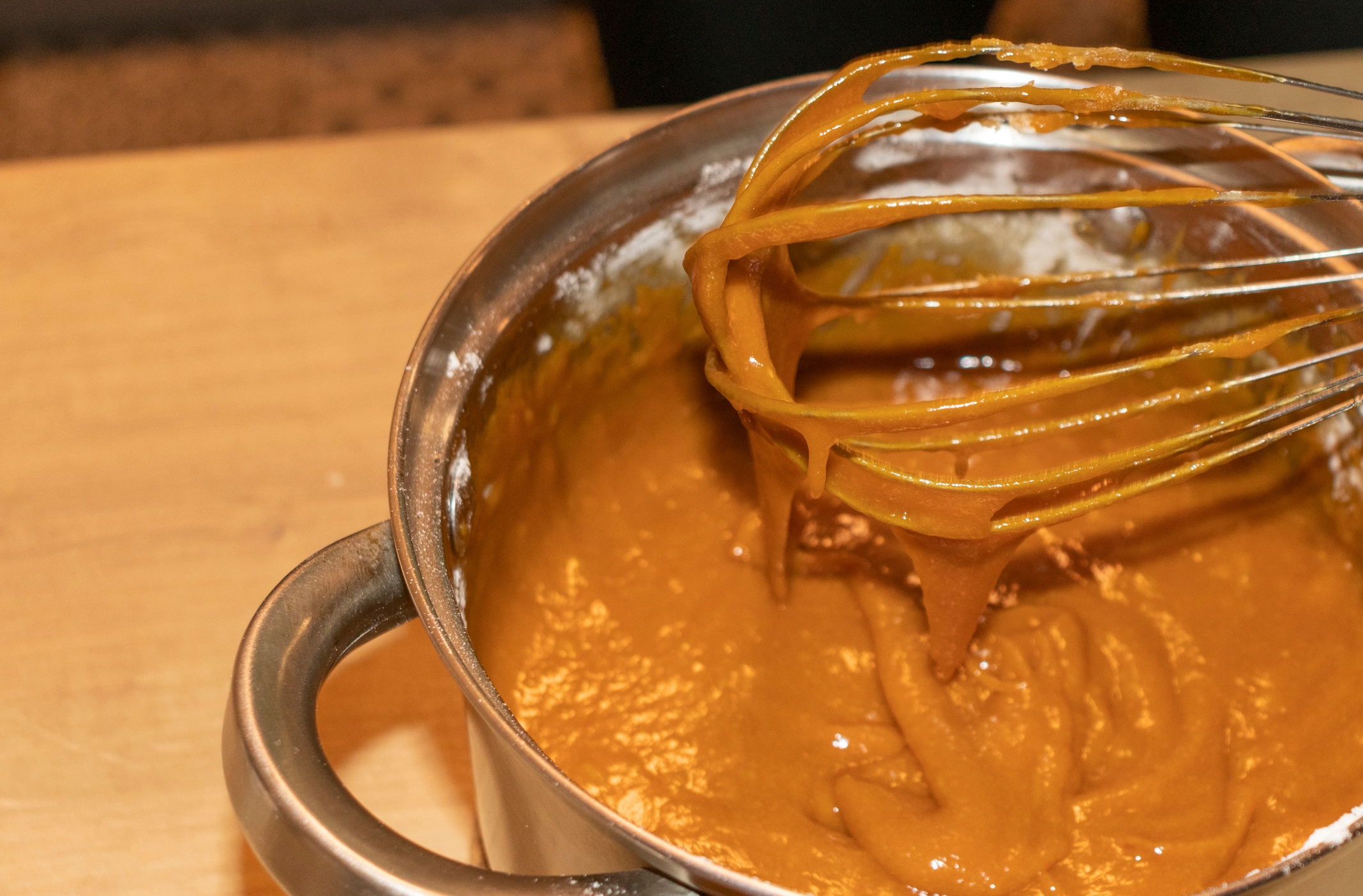 A whisk stirring thick caramel sauce in a metal pot on a wooden surface