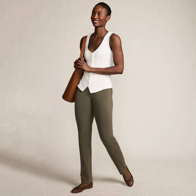 Person smiling in casual attire, wearing a sleeveless top, slim-fit pants, and flats while holding a handbag, posing confidently