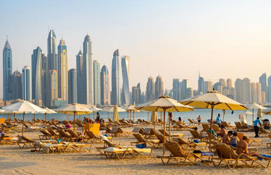 Expatriates sunning themselves on loungers with umbrellas in the evening sun on Fairmont Hotel beach on the Palm, Jumeirah opposite the looming skyscr