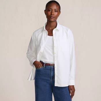 A person models a casual outfit with a white shirt over a tank top and blue jeans, showcasing style for shopping purposes