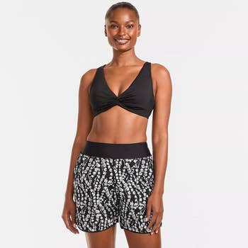Woman in a black twist-front bikini top and patterned swim shorts, smiling at the camera
