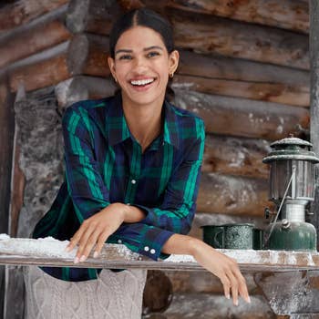 Person smiling, wearing a plaid shirt and cable-knit pants, leaning on a snowy wooden bench with a vintage lantern, in a rustic setting