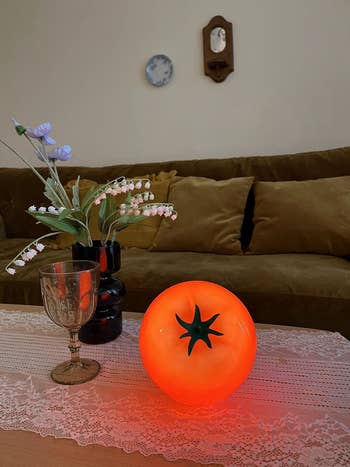 Glowing tomato-shaped lamp on a lace-covered table with flowers, a glass, and a couch in the background