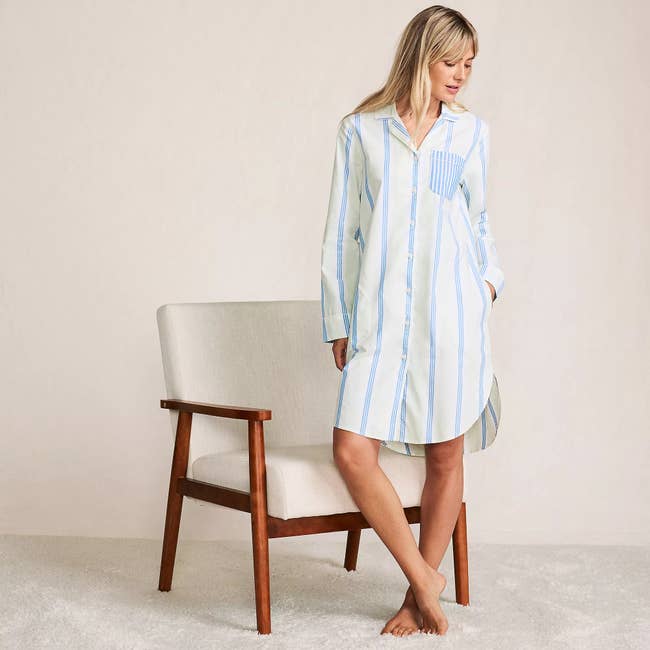 Woman wearing a striped, long-sleeve shirt dress stands beside a modern chair