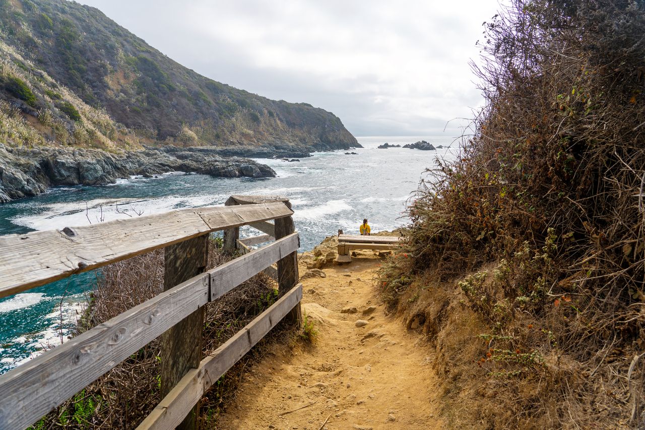 A dirt trail with a wooden railing leads to a cove by the ocean surrounded by hills and cliffs