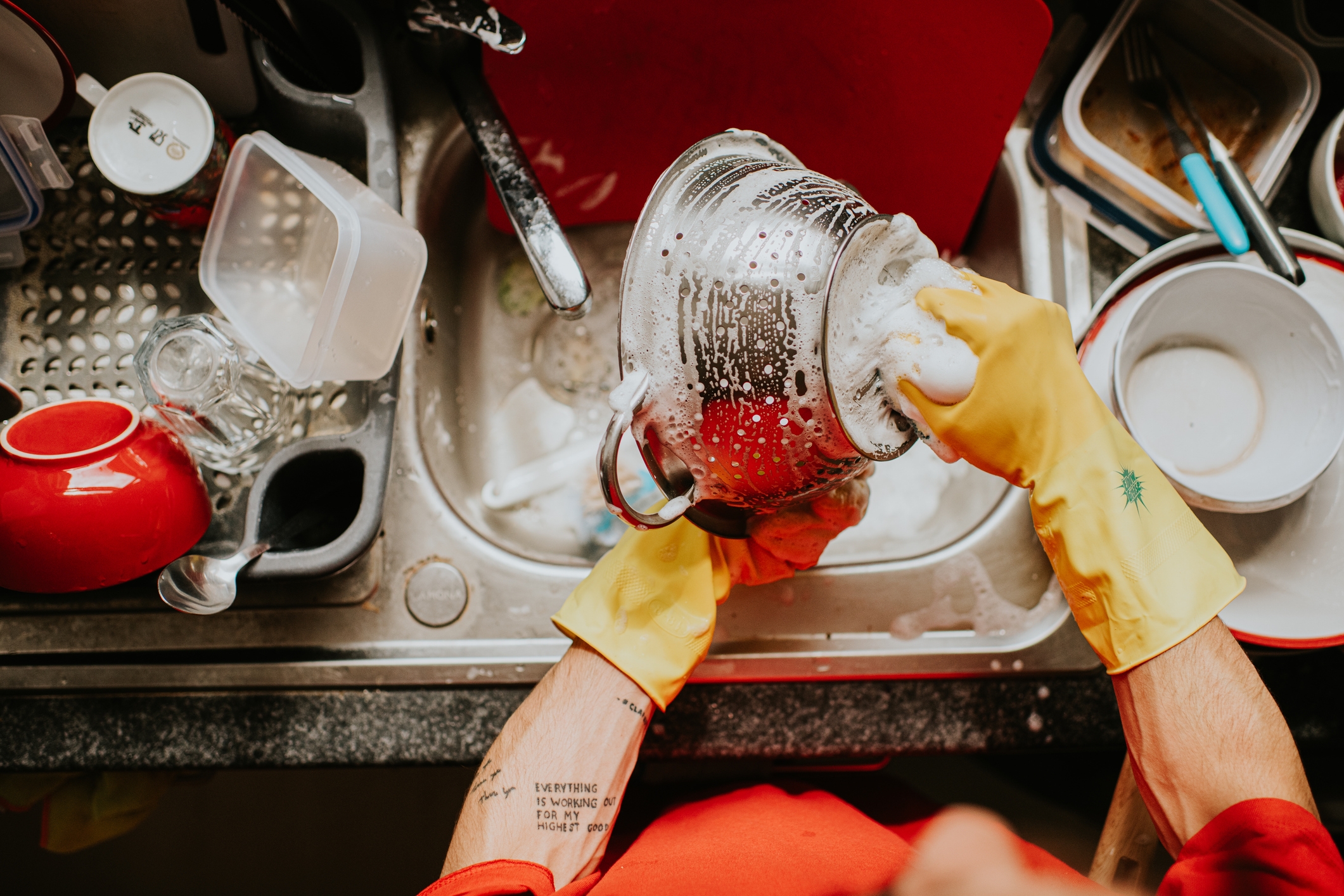Person wearing yellow gloves washing a colander in a kitchen sink full of dishes and foamy water
