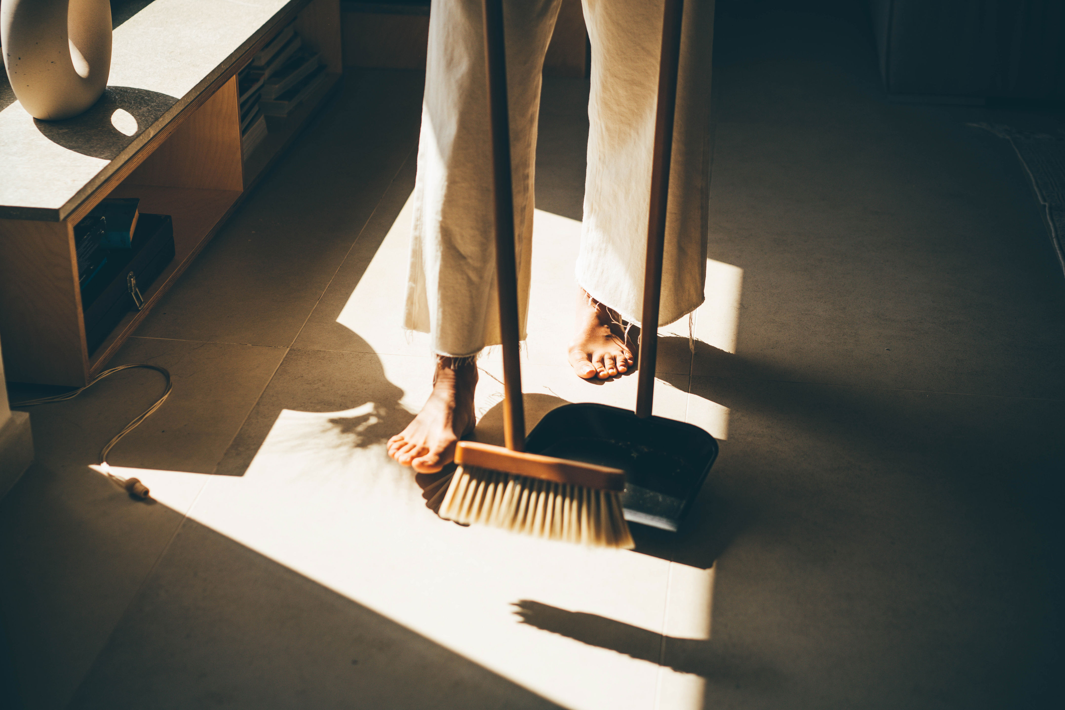 Person sweeping floor with broom and dustpan, sunlight filtering through window