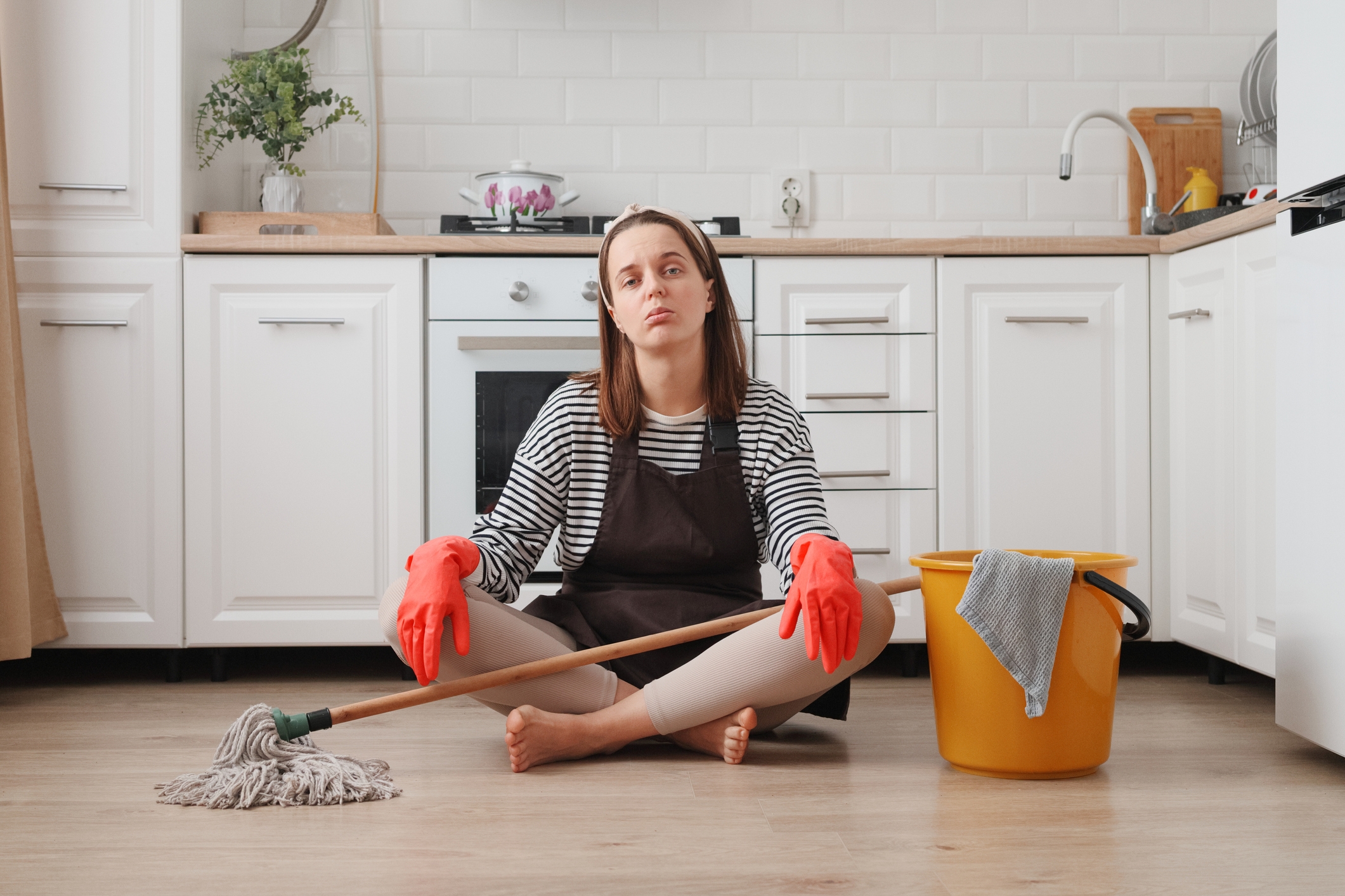 Person sitting on a kitchen floor, wearing cleaning gloves, holding a mop, and looking tired next to a bucket