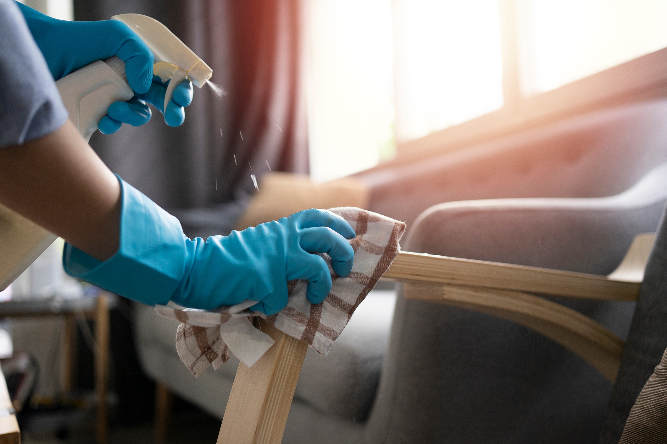 Person cleaning furniture with spray and cloth, wearing blue gloves, in a well-lit room