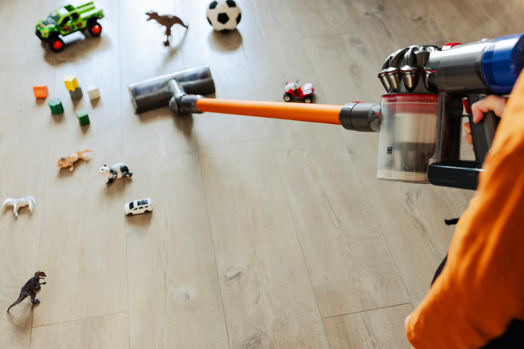 A person vacuuming a wooden floor scattered with toys, including a soccer ball and toy animals