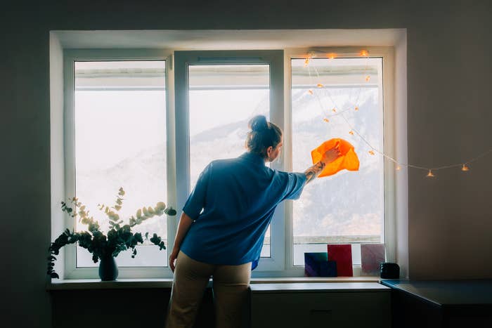 Person cleaning a window with an orange cloth, standing in a room with a vase of flowers and string lights, creating a cozy atmosphere