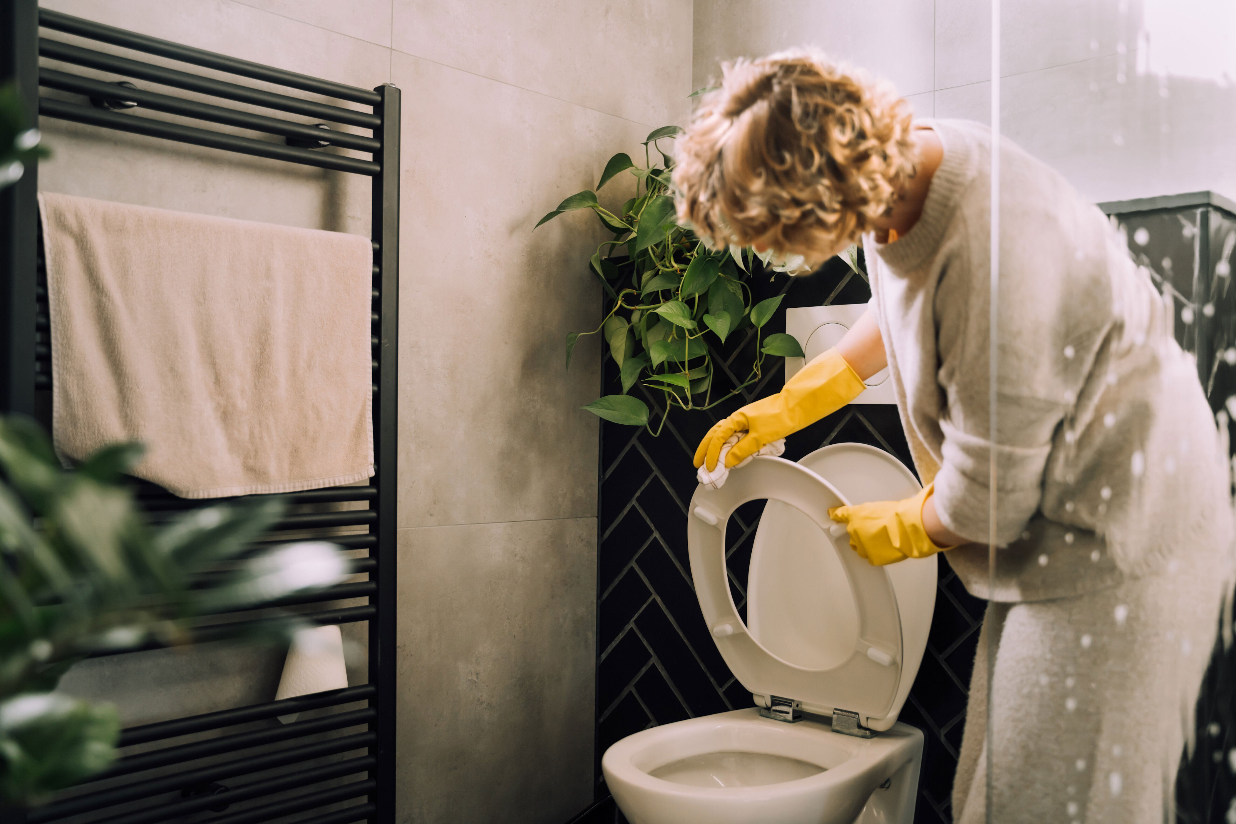 Person in casual clothes cleaning a toilet with gloves on. A towel hangs on a rack, and a plant decorates the bathroom