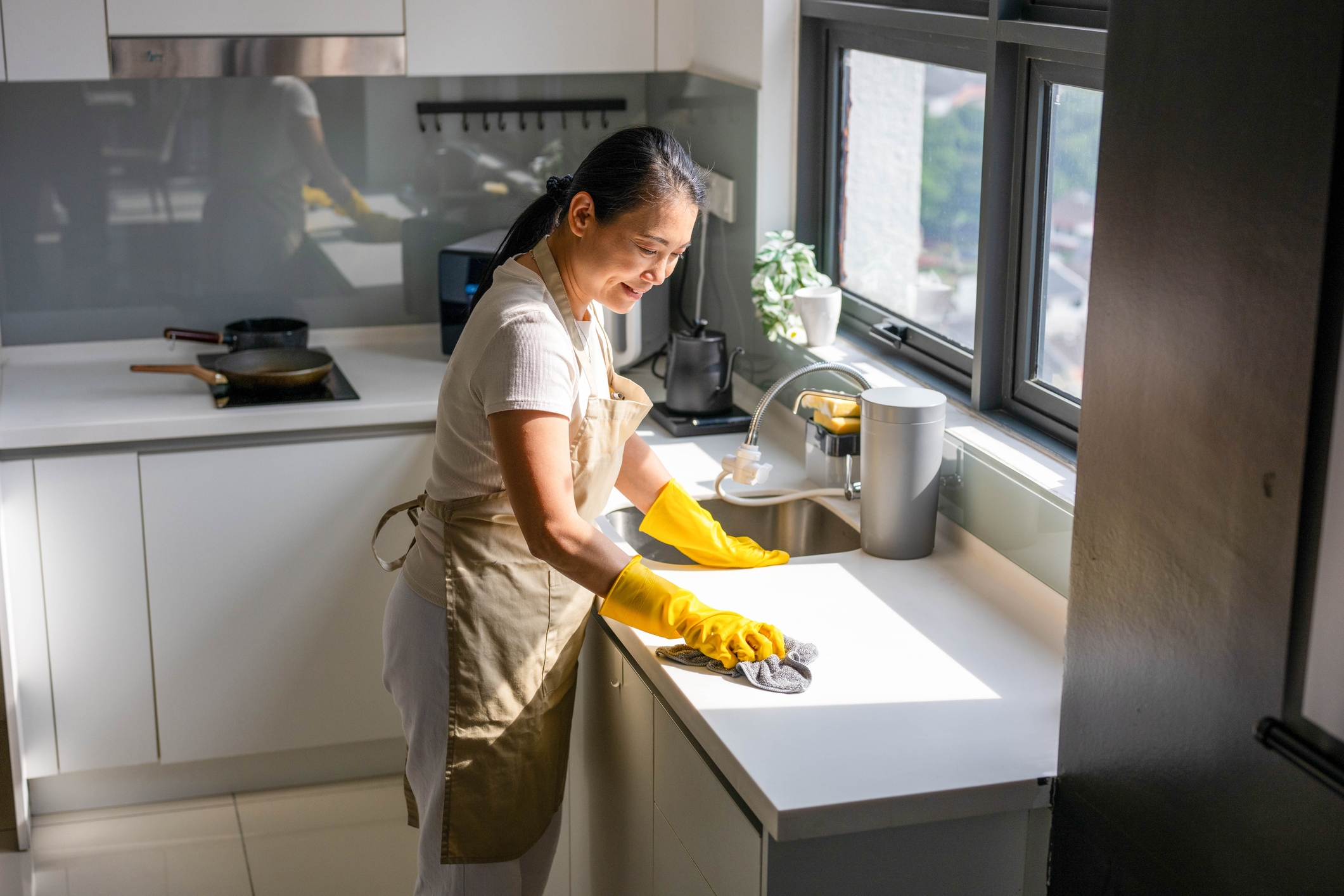 Person cleaning a kitchen counter with gloves, wearing an apron, in a bright kitchen