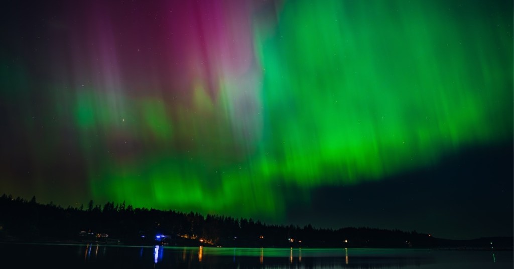 A vivid display of the northern lights casts the night sky in green and red colors above a sleepy forest town on the waterfront.