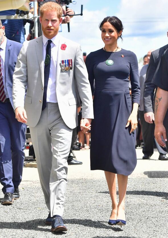 An individual in a white suit with a lapel pin and a tie is walking alongside a person in a dark blue long-sleeve dress, both of