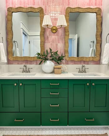 Bathroom with green cabinets, dual sinks, and rattan mirrors. Pink tiled wall backdrop, vase with greenery, and wall-mounted light fixture