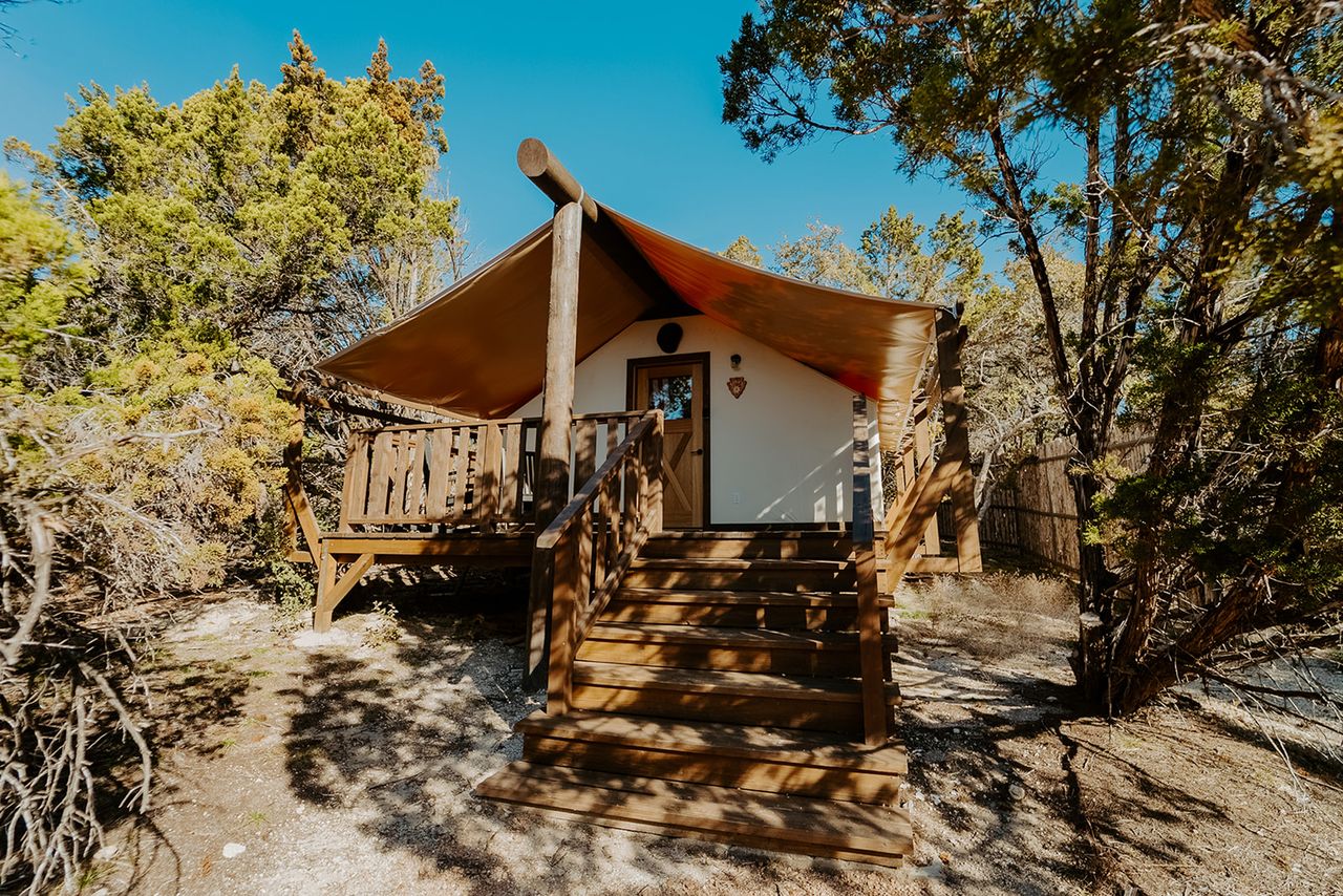 A safaristyle cabin with a canvas awning in a wooded area featuring a front porch and wooden stairs leading up to the entrance
