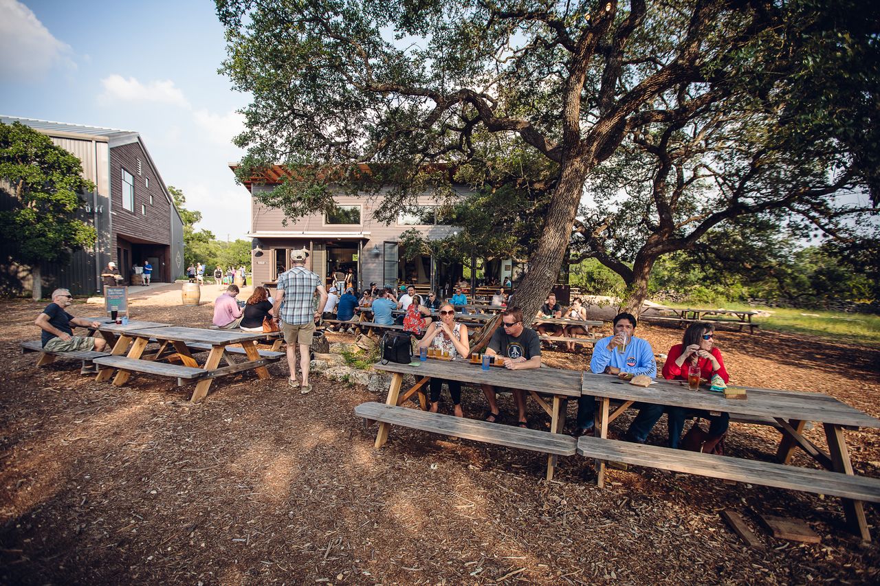 People sitting at outdoor tables under trees in front of a brewery building