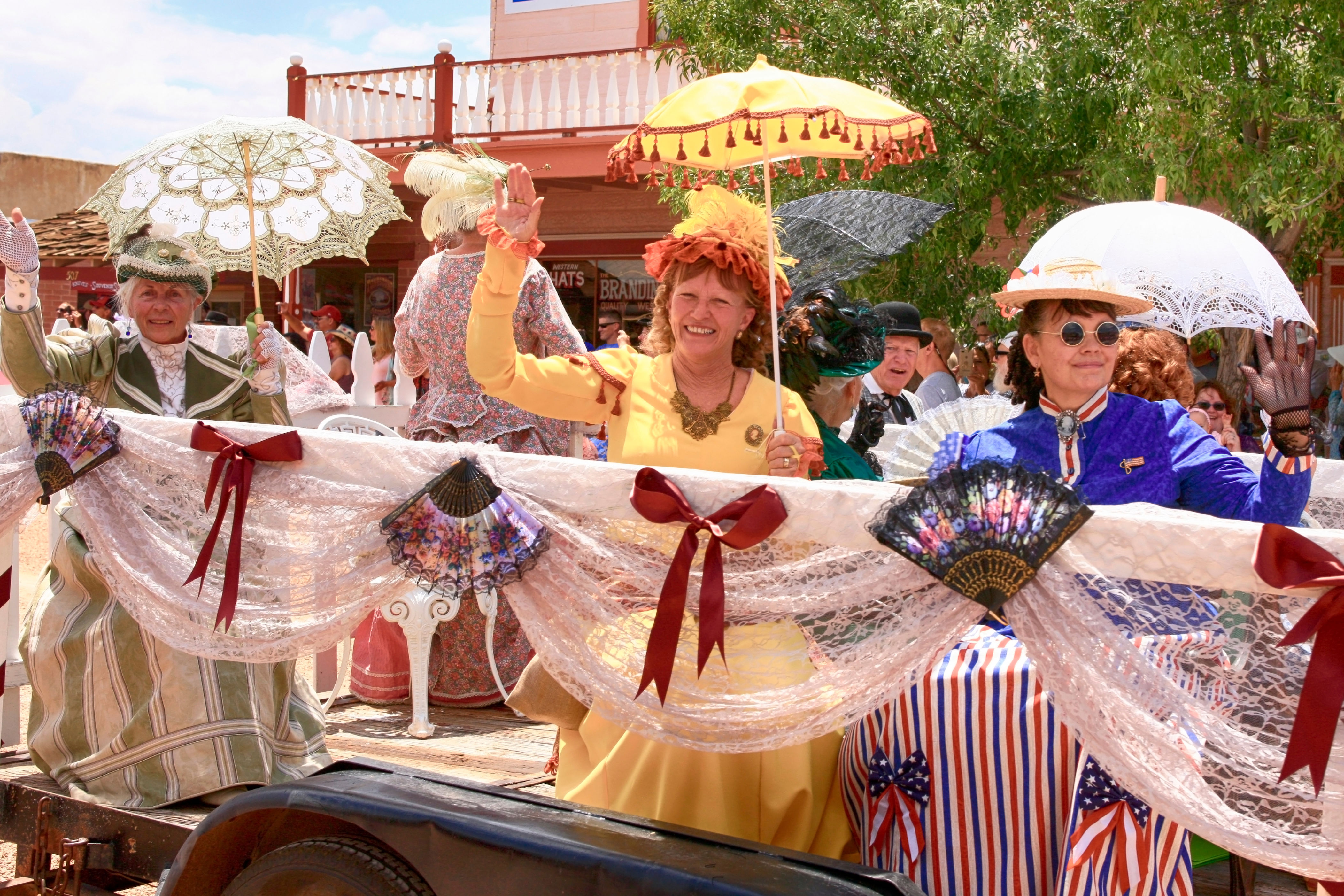 People in vintage-style clothing wave while riding a parade float decorated with lace and fans