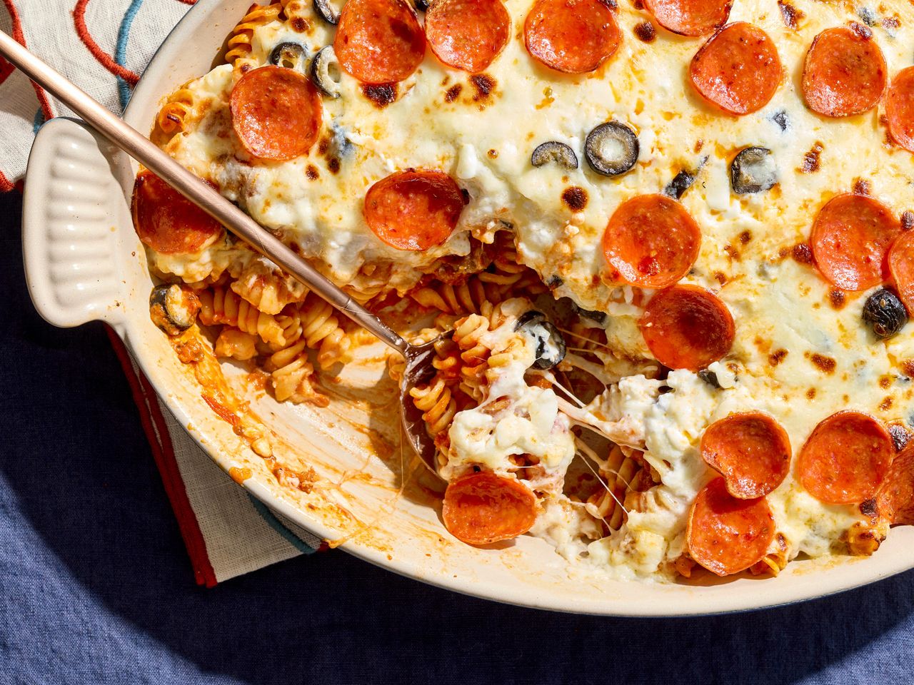 A casserole dish filled with a pepperoni and cheese pasta bake, with a portion removed showing the layers of pasta and sauce