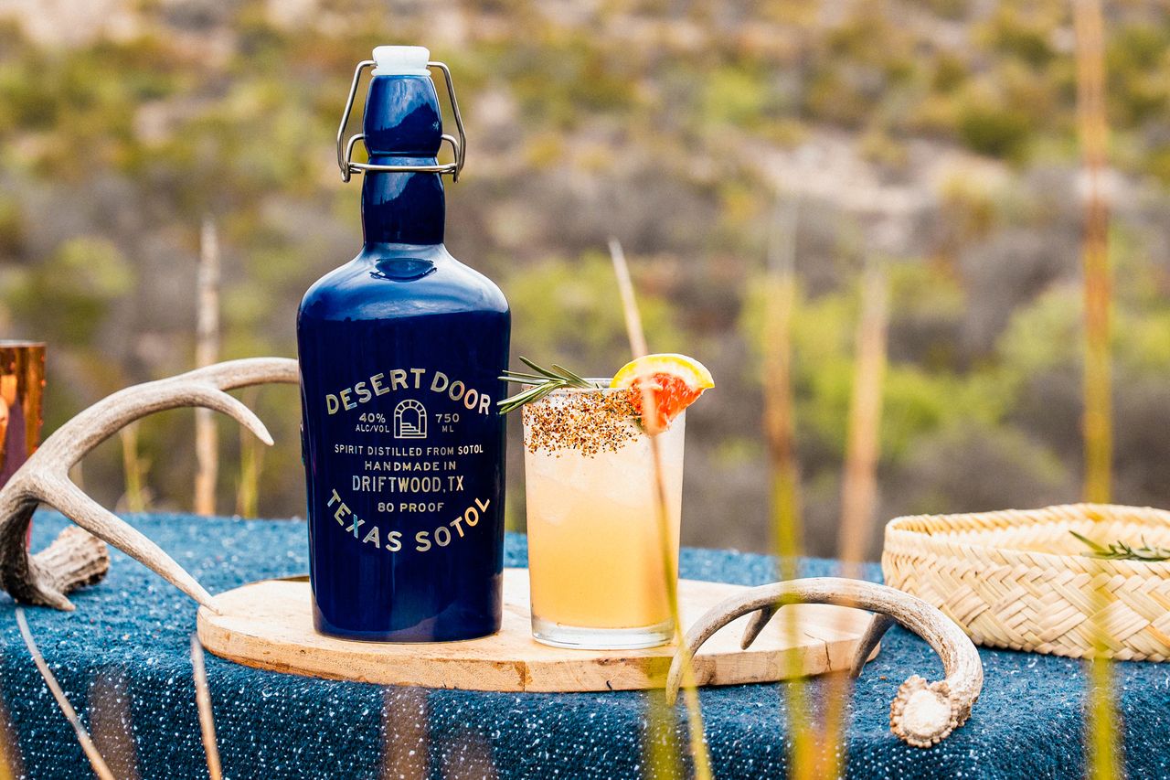 A Desert Door Texas sotol bottle beside a garnished cocktail glass on a wooden serving board outdoors