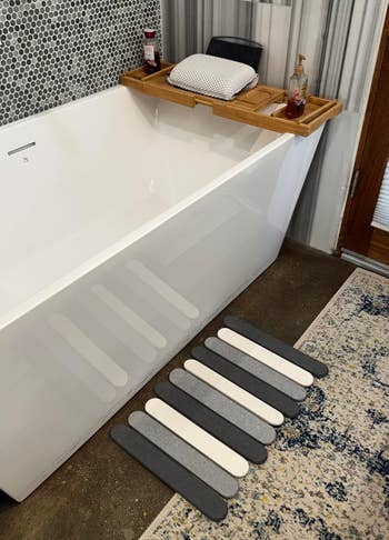Bathtub with a wooden tray holding a pillow and soap, striped wall tiles, and striped bath mat on the floor