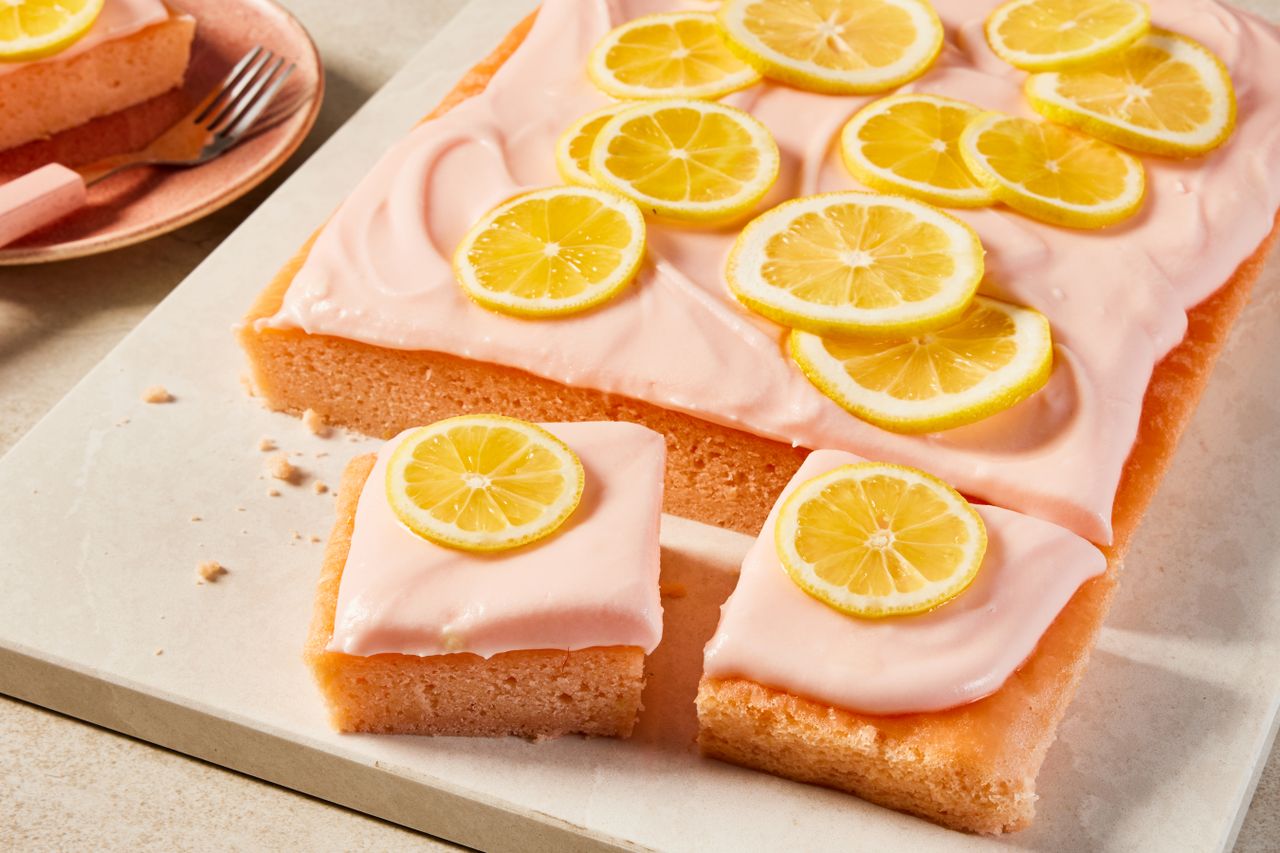pink lemonade cake (with some slices cut out) on a board next to a plate with a slice