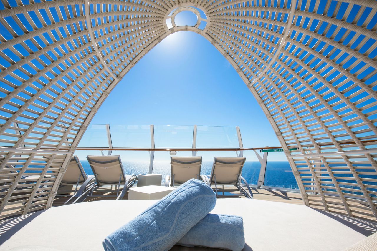 A folded towel rests on a bedlike surface under a lattice dome with lounge chairs and a view of the ocean beyond