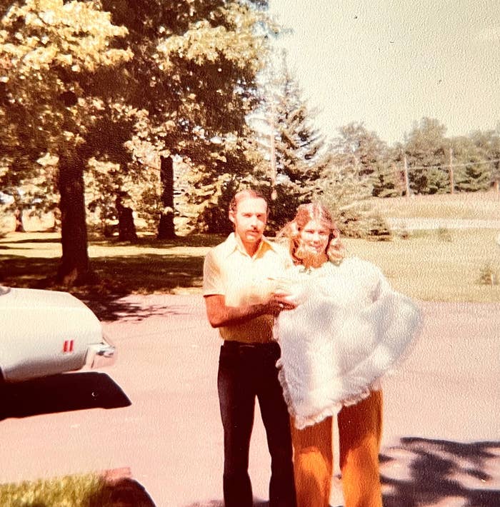 A man and woman stand outside, possibly in the 1970s. The man is in a short-sleeve shirt and pants, the woman in a dress with a flowing shawl. Trees are in the background