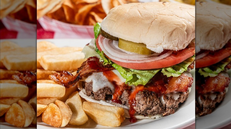 Burger with onions, lettuce, bacon, tomato, and pickl on plate with fries