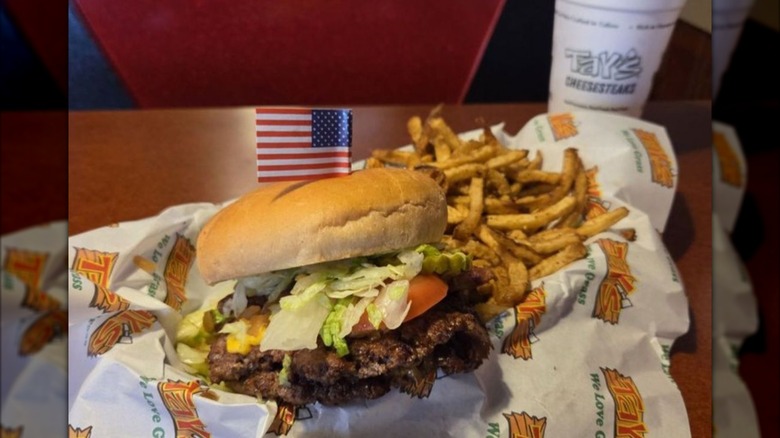 Burger with lettuce and tomato on paper with pile of fries and drink in background