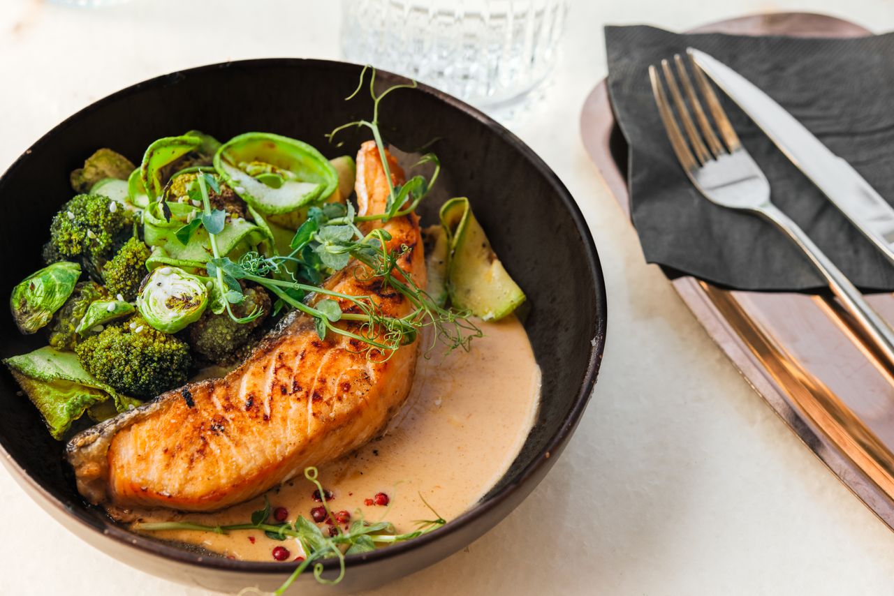 Cooked salmon served in a bowl with vegetables and sauce placed on a table setting with utensils and a napkin nearby