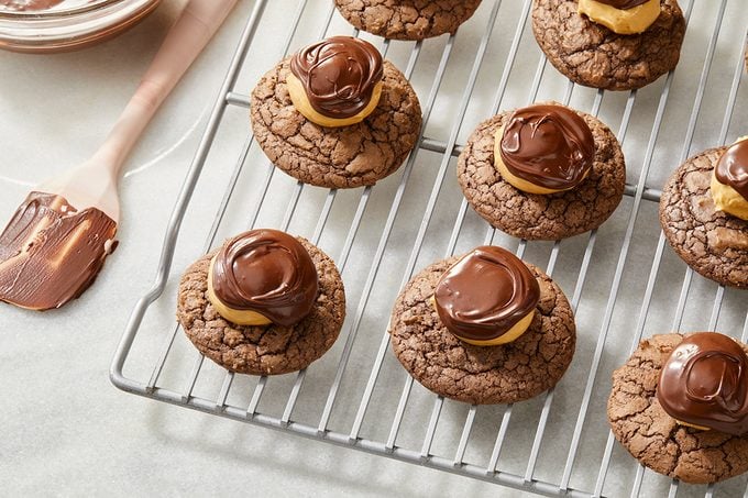Chocolate cookies with peanut butter and swirled chocolate topping are cooling on a wire rack; a spatula with chocolate is nearby on a light countertop.