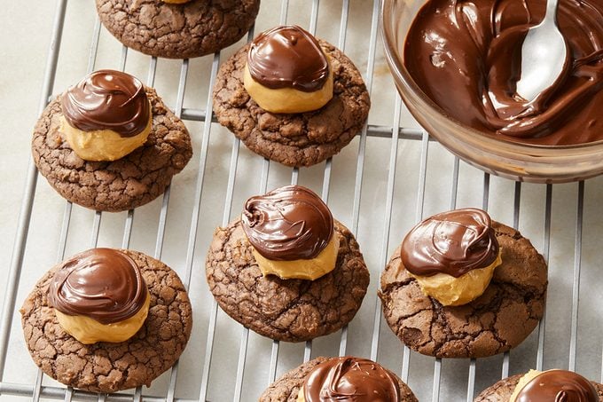 Chocolate cookies topped with a dollop of peanut butter and a swirl of melted chocolate are arranged on a cooling rack. A bowl of melted chocolate with a spoon is beside the cookies.