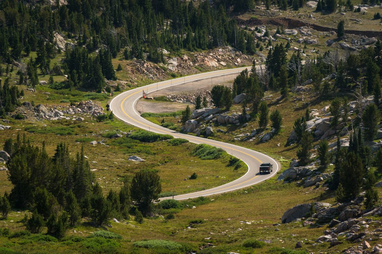 A winding mountain road surrounded by a forest and open fields with a vehicle traveling along the road