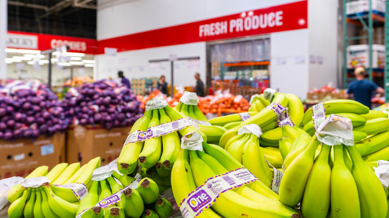 Bananas in a Costco produce section with a large red sign reading "FRESH PRODUCE" in the background.